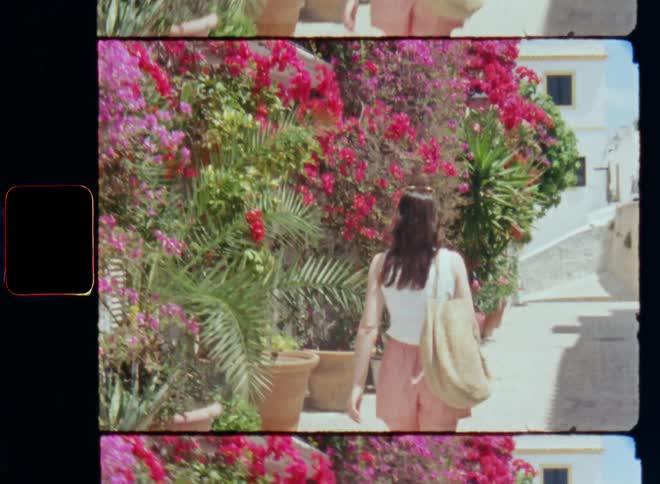 A woman carrying a bag over her shoulder is walking on a small backstreet or alley with loads of flowering green plants and vibrant colourful flowers on the side of the road next to the homes and growing on the stone walls on nostalgic film