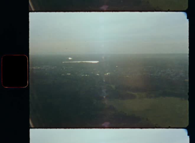 Flying over a populated rural area with a sprawling city surrounded by farmland on open fields and green forests with a lake or large body of water in the background at the horizon while the sun is shining brightly