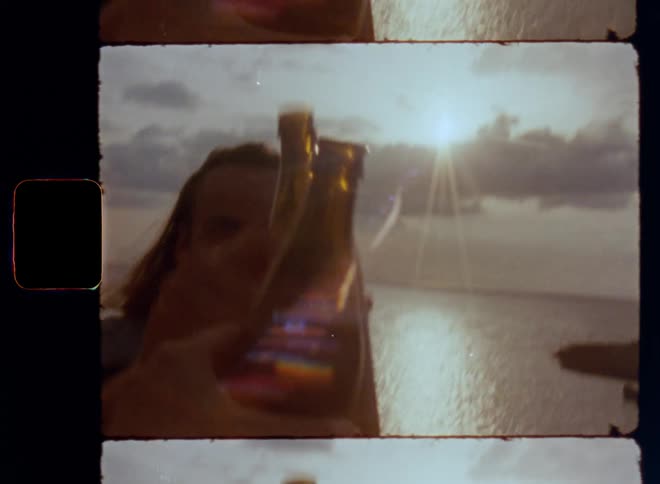 POV Super 8mm film shot of a hand holding a bottle that taps against a man’s beer in a celebratory tone while celebrating life and toasting to good times with happy and careless summer vibes as he smiles expressing happiness and enjoyment in the sun