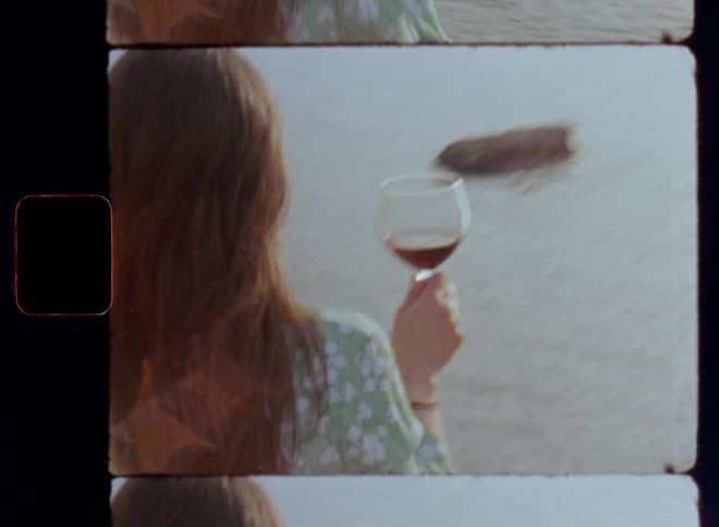 Dynamic handheld 8mm film clip of a brunette woman in a green flowered summer dress standing on a ridge overlooking the ocean while holding a wineglass filled with red wine in her hand as she looks into the bright sunlight of the setting sun that shines