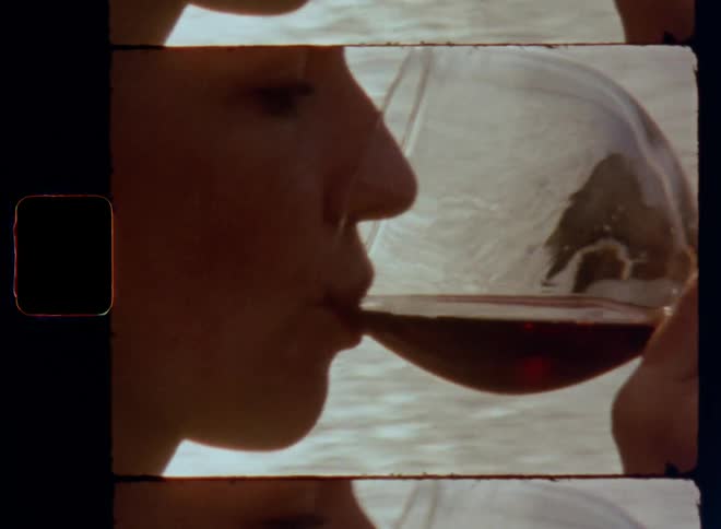 Super-8 close up of the mouth of a woman that drinks flavorful red wine from goblet glass enjoying the great taste stimulating her gustatory sense with a tasteful sensory impression near ocean or lake with sunshine backlight from the rear