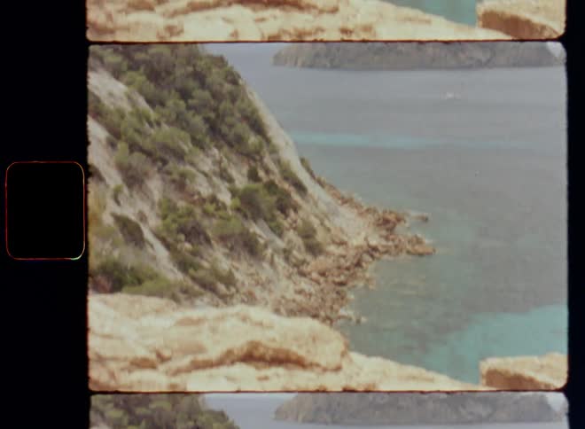 Cracks in a dangerous and steep rocky seaside cliff in front of a small island with high rocky mountain formation on a cloudy day with overcast sky in the ocean with blue salt water on old Super-8 Film footage with tilting camera movement