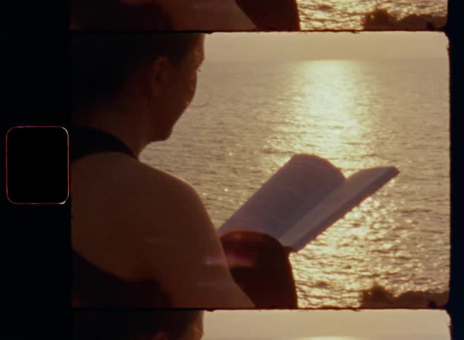 Super 8mm close up of a woman holding a book in her hands while reading on the beach as the sun rises at the horizon shining onto her skin warming the fresh air by the sea and reflecting on the surface of the water causing lens flare and glare
