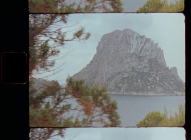 Super 8 film footage of a wooded area revealing the view onto a mountainous landscape rising to the overcast sky with cloud cover in the ocean surrounded by blue sea water with steep cliffs and rocks on the shoreline