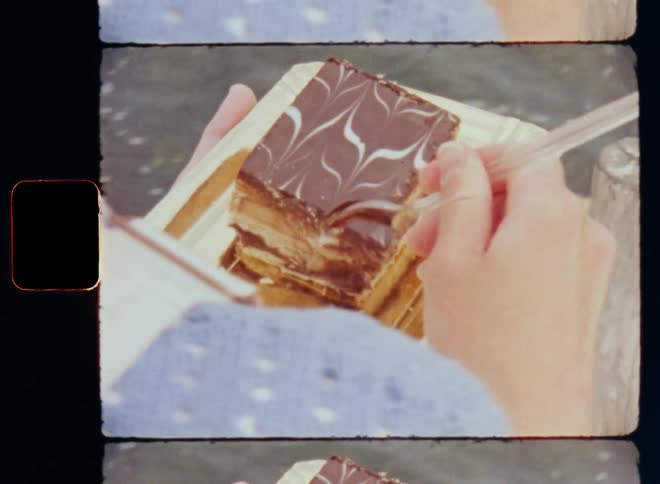 Close up of a woman in a knitted blue sweater eating delicious and sweet chocolate cake off a golden plate using plastic spoon while she sits on a pier or harbor wall by the water of the sea or ocean enjoying the confectionery treat