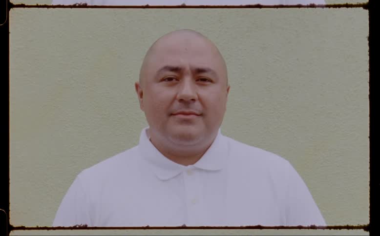 Face of a bald man wearing a white shirt standing in front of a wall