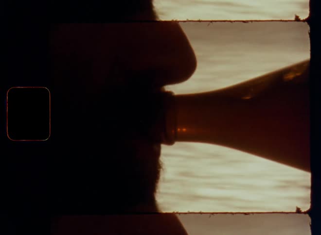 Closeup of a bearded man with short beard drinking or sipping a sip from brown glass bottle filled with a refreshing beverage or cold beer illuminated with sunshine from the back for great refreshment on a warm summer day at the beach during vacation