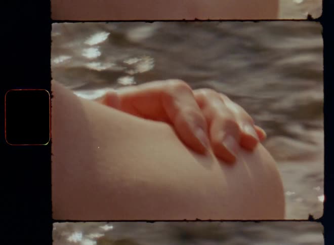 A person or young woman places a hand on her back touching her soft naked skin expressing self-care and self love while sitting in the shallow water at the beach as the sun shines and warms up the bay at the ocean lake or sea on Super-8 film