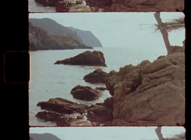 Close up of coastal rocks and cliffs in a sheltered bay by the sea or ocean as the sun shines an reflects the water during the day with nice weather with handheld camera movement and high steep mountains in the far distant background