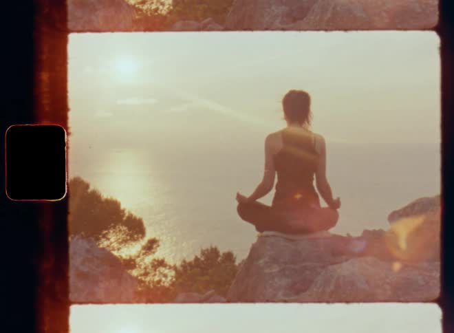 Slowly panning Super 8mm shot of young woman in black top and leggings sitting on high rock cliff and doing yoga or meditating at sunset watching the sun go down at the horizon high above the sea relaxing for body awareness and stress relief