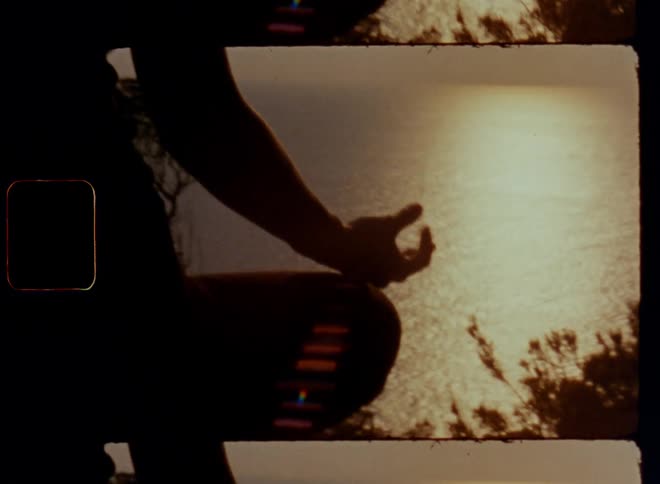 Low angle sunshine shines onto a young woman during yoga or meditation session while sitting on a rock on a mountain or cliff overviewing the ocean or sea during sunset on a warm summer day while laying her hand onto her leg and enjoying quietness