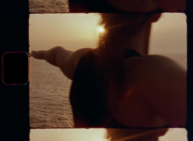 Dynamic handheld 8mm film of muscular well-trained woman with brown hair doing a warrior yoga pose as symbol for strength energy and power with both arms stretched sideways at sunset by the beach with light reflection on the ocean or sea water