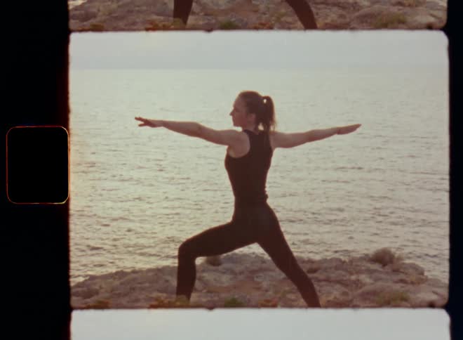 Brunette woman with long hair and black sports apparel stretching and raising her trained arms doing yoga or Pilates exercises by the sea in front of the ocean while light breeze or wind ripples the surface causing waves and movement and sun reflection