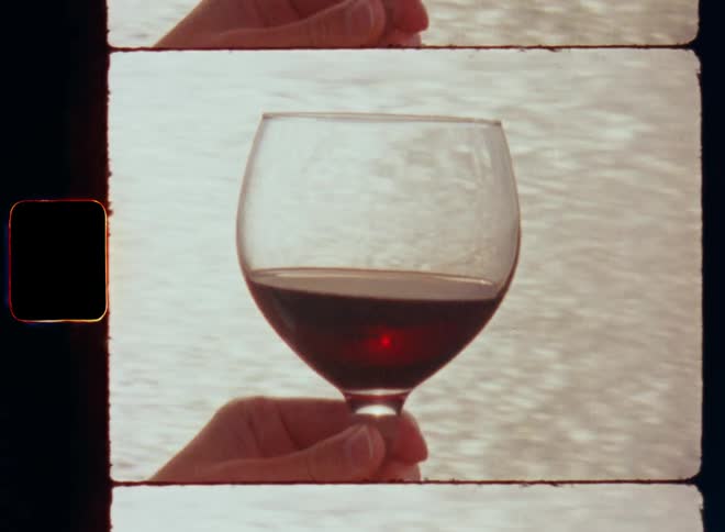 Someone is holding a transparent glass of red wine in his or her hand in front of the white or bright water surface of a lake river or the sea as the camera moves sideways and light reflects on the ocean and the liquid is glowing or glooming in the sun