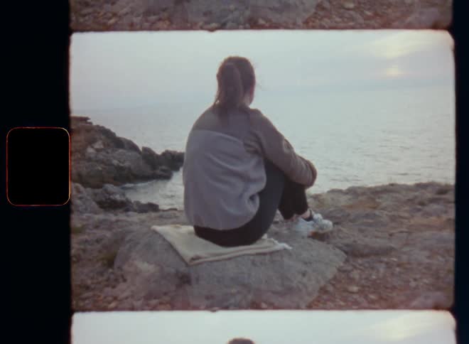 Brunette woman wearing retro fleece pullover and sneakers sitting on a rock staring or looking at the horizon at sea in a contemplative and thoughtful way while thinking about life and enjoying the quiet and peaceful atmosphere on Super-8 vintage film