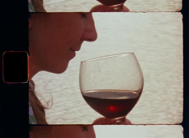 Close up woman holding glass of red wine in her hand as she lifts it to her nose and smells intense flavor in front of the ocean or sea with a bright glare or reflection of the setting sun during evening time in the background on Super 8mm film footage