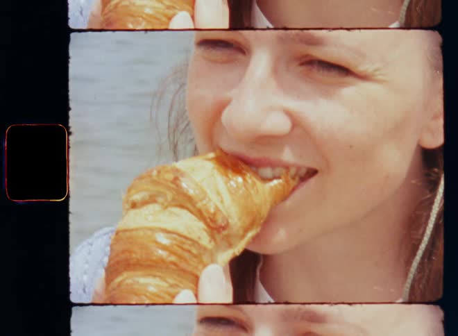 Close up of a woman in a blue knit sweater eating a freshly baked and warm crescent roll or croissant during the day near water in warm sunlight and she smiles happily after she bites into the delicious pastry enjoying the satisfaction of consuming sugar