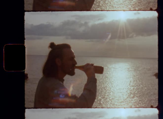 Young man raises beer bottle while saying cheers and drinks a sip after toasting and smiling to friends while standing on a cliff by the ocean enjoying a mild spring evening in great company on retro Super 8mm film with bright sunshine and lens flare