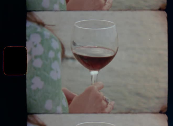 A woman is drinking red wine by the water or beach of the sea as she holds the glass in her hands with an expensive silver jewellery ring representing wealth and richness on Super-8 with smooth sideways camera movement