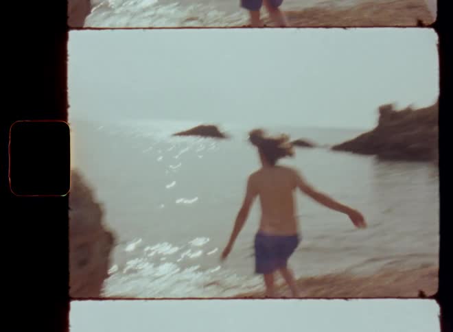 Young man takes off his white shirt as he runs or walks towards water edge on the beach in bay before he stops at the edge of the ocean or sea and looks at the flickering surface with sun light reflections in front of horizon feeling free and happy