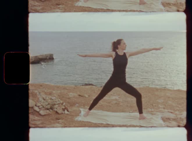 Handheld dynamic 8mm film footage of a young and fit woman symbolizing mobility and flexibility who raises her arms in yoga movements with a forward and a backward bend at the shoreline of a large lake or the sea with the sun going down at the horizon