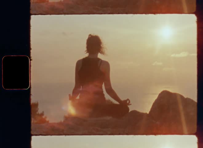 A woman sitting Indian style with crossed legs while meditating during a relaxing zen yoga session facing the ocean as the sun goes down at the horizon and warm sunrays shine onto her body connected to her mind and the hair sways in the breeze
