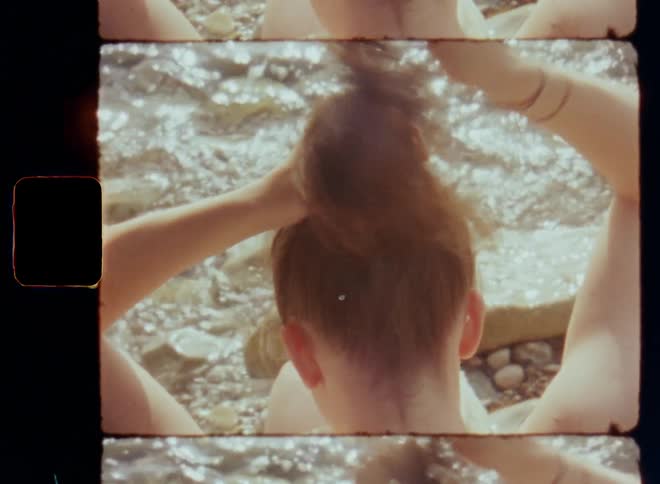 Close up of lightly dressed brunette woman tying her long brown hair to a ponytail using a hair tie or scrunchy for a bun or chignon hairstyle while sitting at the beach next to the moving water in the sunshine on vintage super 8mm with retro film burn