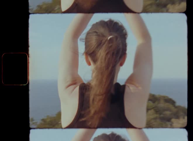Dynamic handheld 8mm film walking towards a sportive lady doing exterior Pilates or outdoor yoga on a cliff with high elevation and great view over the sea or ocean as she puts both palms of her hands together close to the chest while standing