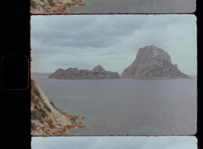 Slowly panning Super 8mm film footage shot of a stunning and spectacular landscape of rock mountains called Es Vedra on Ibiza in Spain in the middle of the Mediterranean Sea with dark clouds above the hill in the ocean