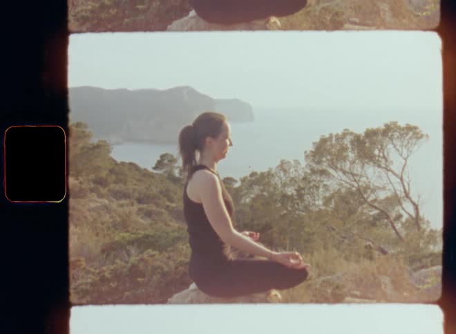 A woman sitting on a rock or large stone doing slow zen yoga or meditation in the middle of nature resembling India or another spiritual country with cliffs hills and mountains at the high coast providing great view over the ocean and the sea on 8mm