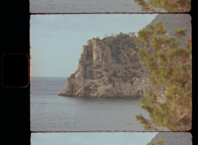 Promontory or rocky cliff on the coast next to the ocean caught on vintage film revealing the coastline behind a green pine tree bush vegetation in wild and pristine nature on overcast and gloomy day with horizontal Super-8 camera movement