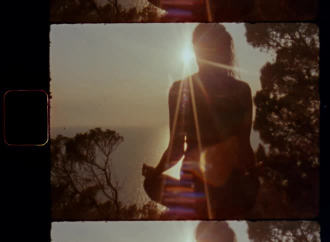 Backside of an inspired woman sitting Indian style with crossed legs while meditating overlooking the ocean as the sun rises in the sky in the early quite peaceful morning hours with golden light on a beautiful summer day in nature outdoors