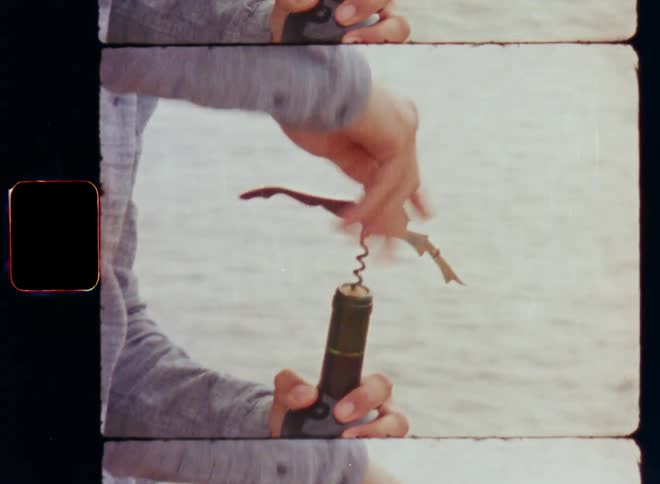 A person wearing a grey shirt with long sleeves opening a green red wine bottle by screwing an opener into the cork while standing at the ocean or sea as the waves move the water surface on vintage Super 8mm film footage