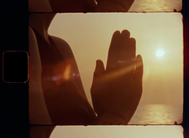 Dynamic handheld close up of a woman meditating with her hands together at her chest or breast standing or sitting at large body of water with the sunlight from the rear shining on the beach as sun sets in the evening at the horizon