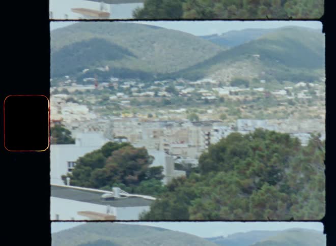 Wide view onto the outskirts of a village or town with man buildings and houses in a valley or vale at the base of mountains covered in green vegetation and trees during the day as the daylight sun shines onto the homes of the city on S8 film footage