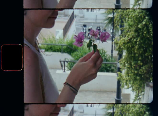 A brunette woman with a white shirt reaches into a flowering bush or tree on the side of the road and picks a branch with green leaves and pink purple or lilac flowers with her hands and smells the fresh scent of spring before she smiles on 8mm film