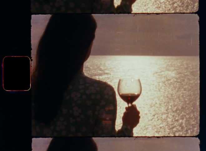 Unrecognisable brunette woman with long brown hair wearing a green flowered dress holds a glass of red wine in her hands as she looks out over the ocean sea as the sun sets at the horizon and clouds cover the sky on vintage film footage