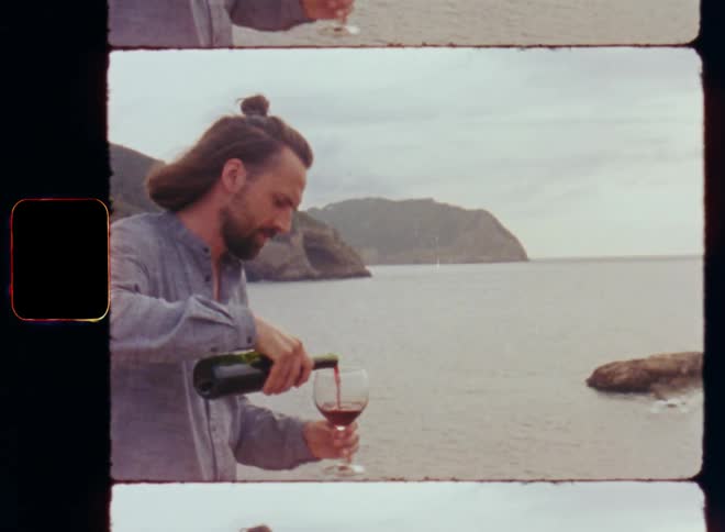 Handheld dynamic 8mm film footage of a man or wine connoisseur in gray shirt pouring a glass of red wine while standing on the beach at the ocean or Mediterranean Sea enjoying the local specialty bought locally