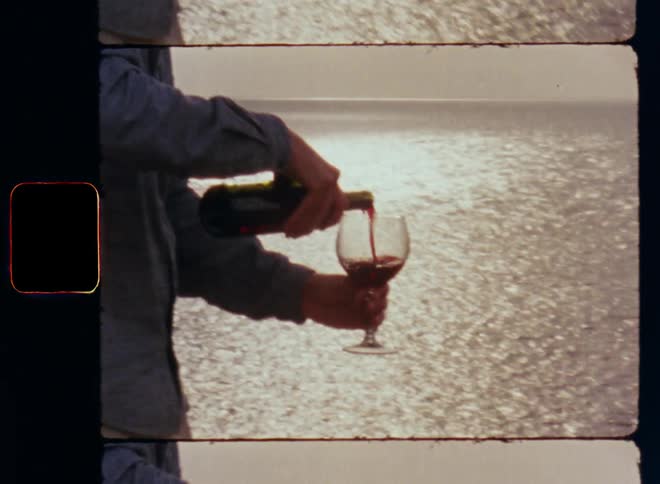 Man fills glass with dry red wine from green bottle before he lifts the beverage to smell the flavor rising from the goblet as the sun sets and reflects on the ripely water surface of a lake sea or ocean while standing at the beach during evening time