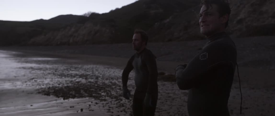 Two men stand on the beach wearing wet suits