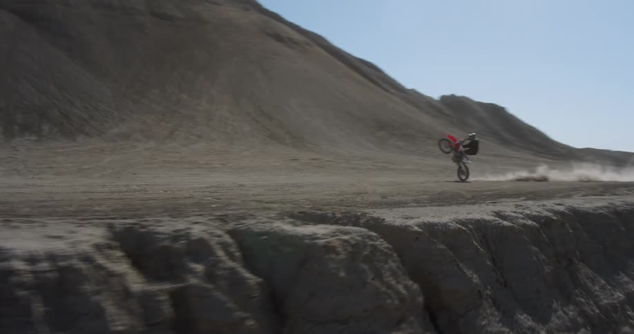 Drone view of a person riding a dirt bike doing a wheelie across desert land near a mountain