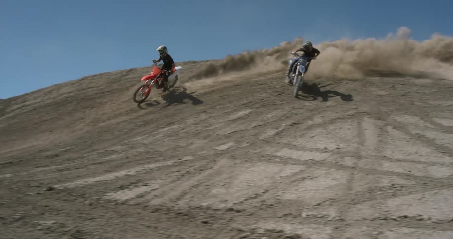 Two individuals riding dirt bikes down a hill fully geared up creating swirling dust in their wake