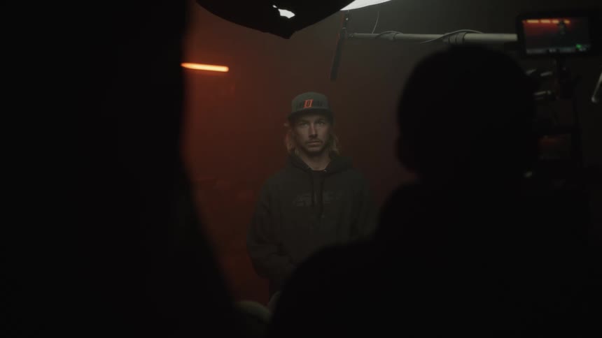 A blond man wearing a cap standing in a dimly lit indoor environment being filmed