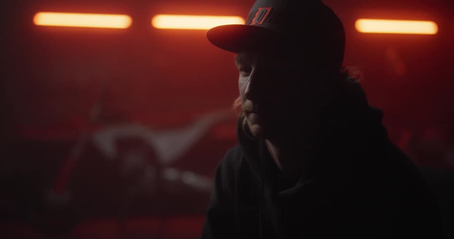 A man in a baseball cap staring straight ahead in thought and a motorbike behind in the background