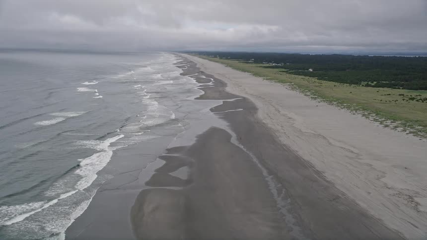 Aerial footage captures the beautiful coastline of Oceanside Washington with ocean waves and a lone person enjoying a leisurely walk along the beach on a cloudy day during summer