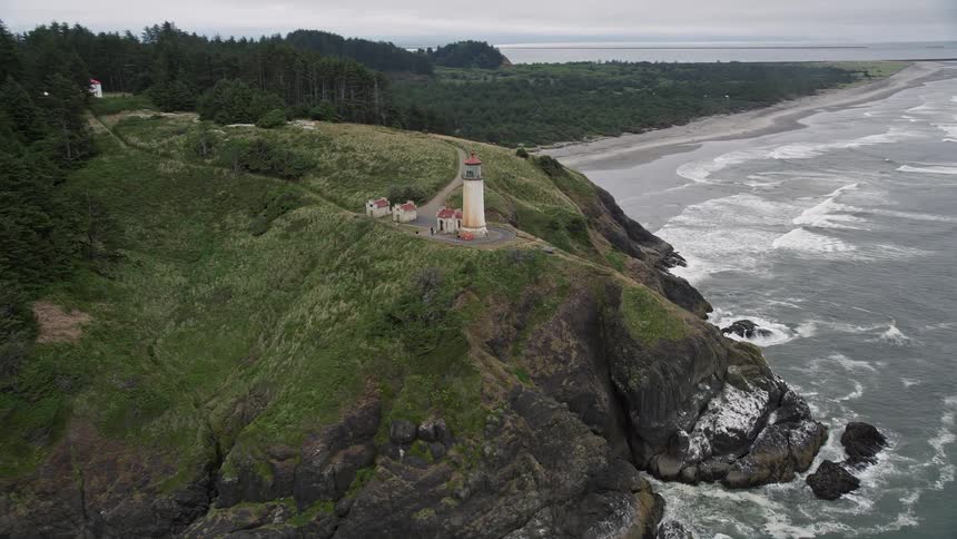 Aerial footage of North Head Light on a coastal cliff with waves crashing against the rocks at the base in Ilwaco Washington during the summer