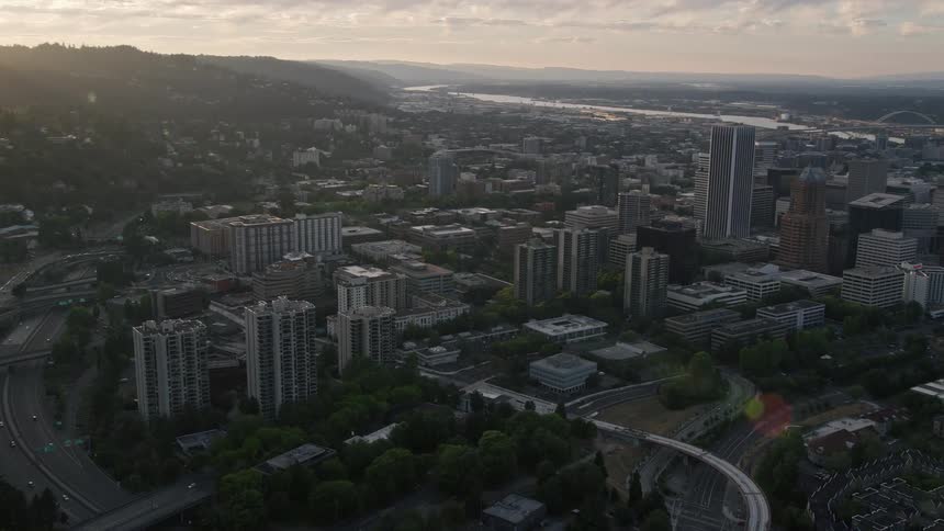 Aerial footage captures the stunning sunset over Portland Oregon as it follows the Willamette River towards the iconic White Stag Sign in downtown showcasing the city's historical landmarks high-rise buildings and the natural beauty of the Pacific Northwe
