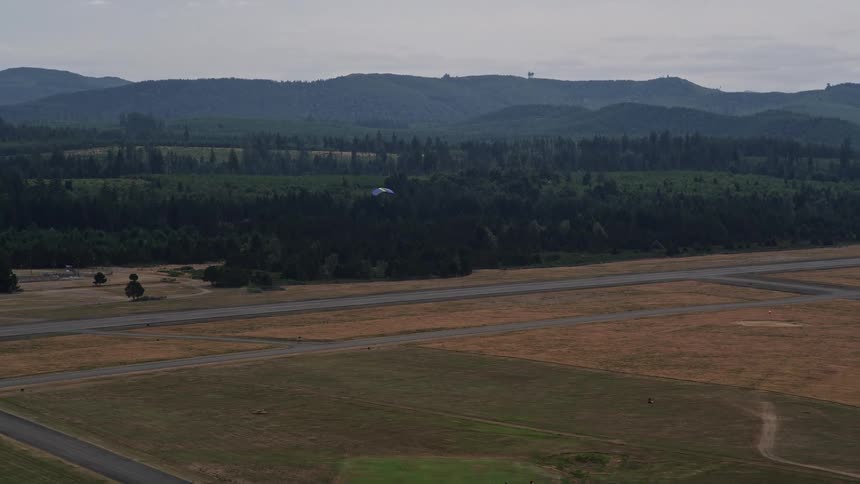 A skydiver gracefully lands on an airfield runway surrounded by parked airplanes and green vegetation at Sanderson Field in Shelton Washington during the daytime in summer