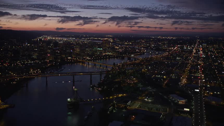 Stunning aerial footage at twilight captures the vibrant downtown Portland skyline the flowing Willamette River and the iconic Ross Island Bridge showcasing the bustling and picturesque cityscape with its high-rise buildings and transportation networks