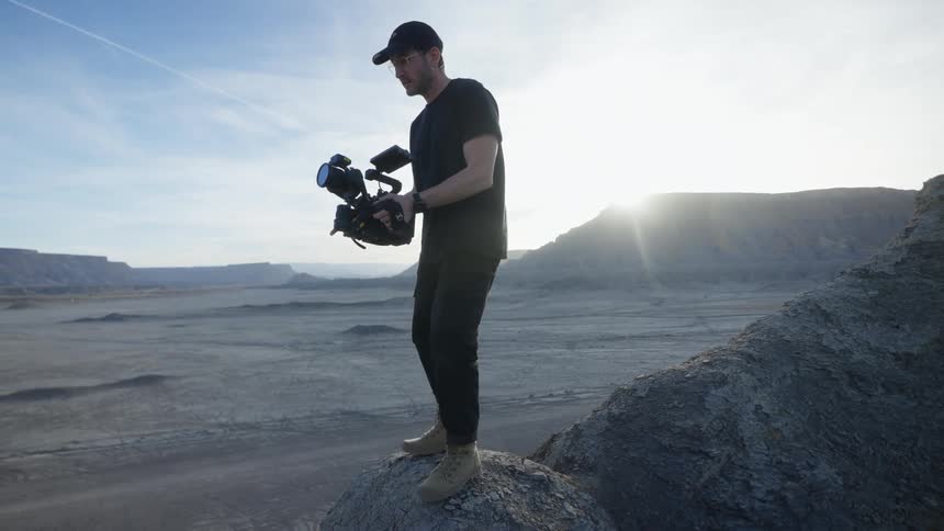 A man on top of a rock in the desert using his camera to film something